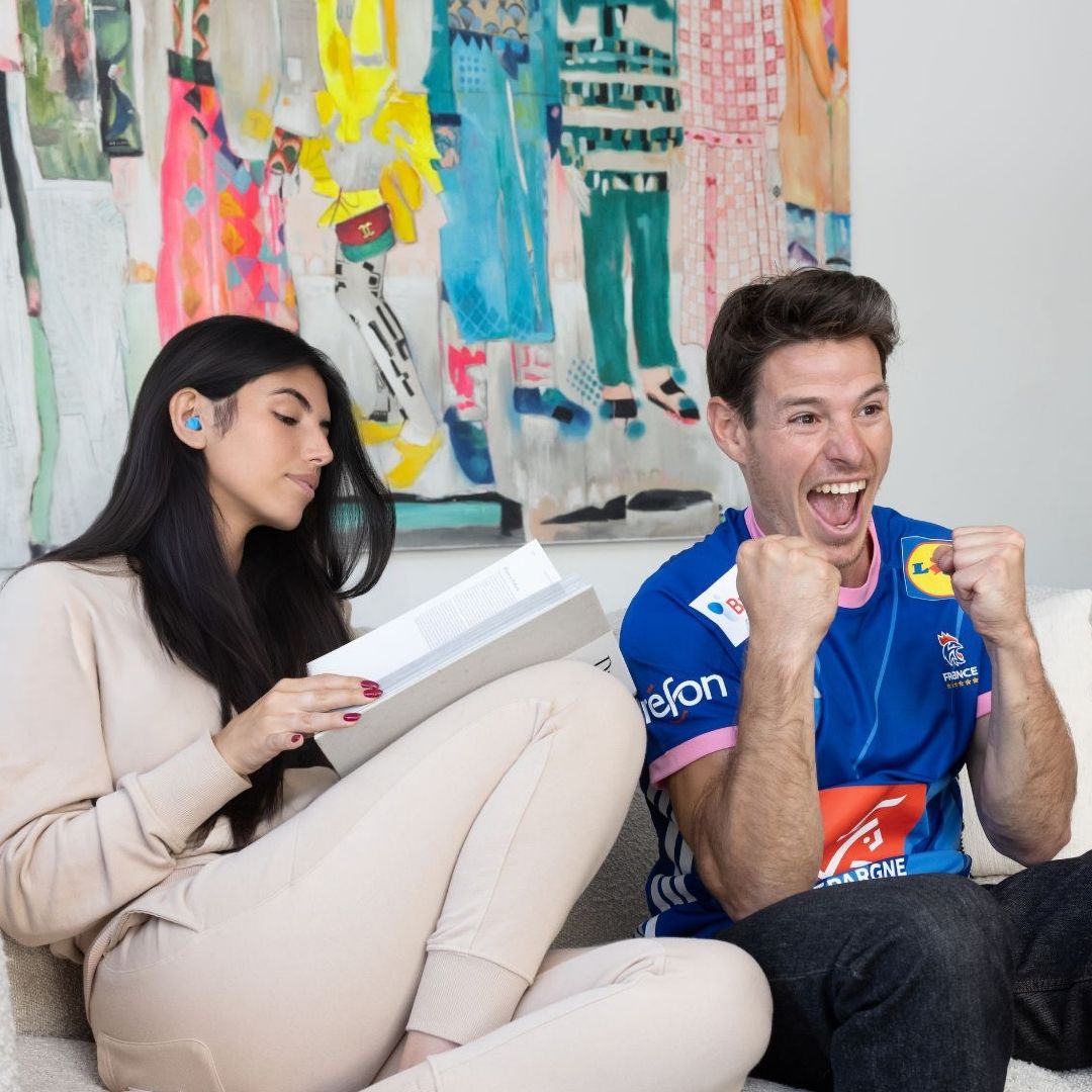 Homme regardant un match à la télévision avec sa femme lisant avec des bouchons Quies Mousse Disco