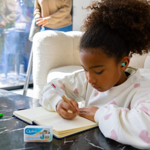 Petite fille faisant ses devoirs avec des bouchons d'oreilles Quies Mousse Mini pendant que sa mère passe l'aspirateur