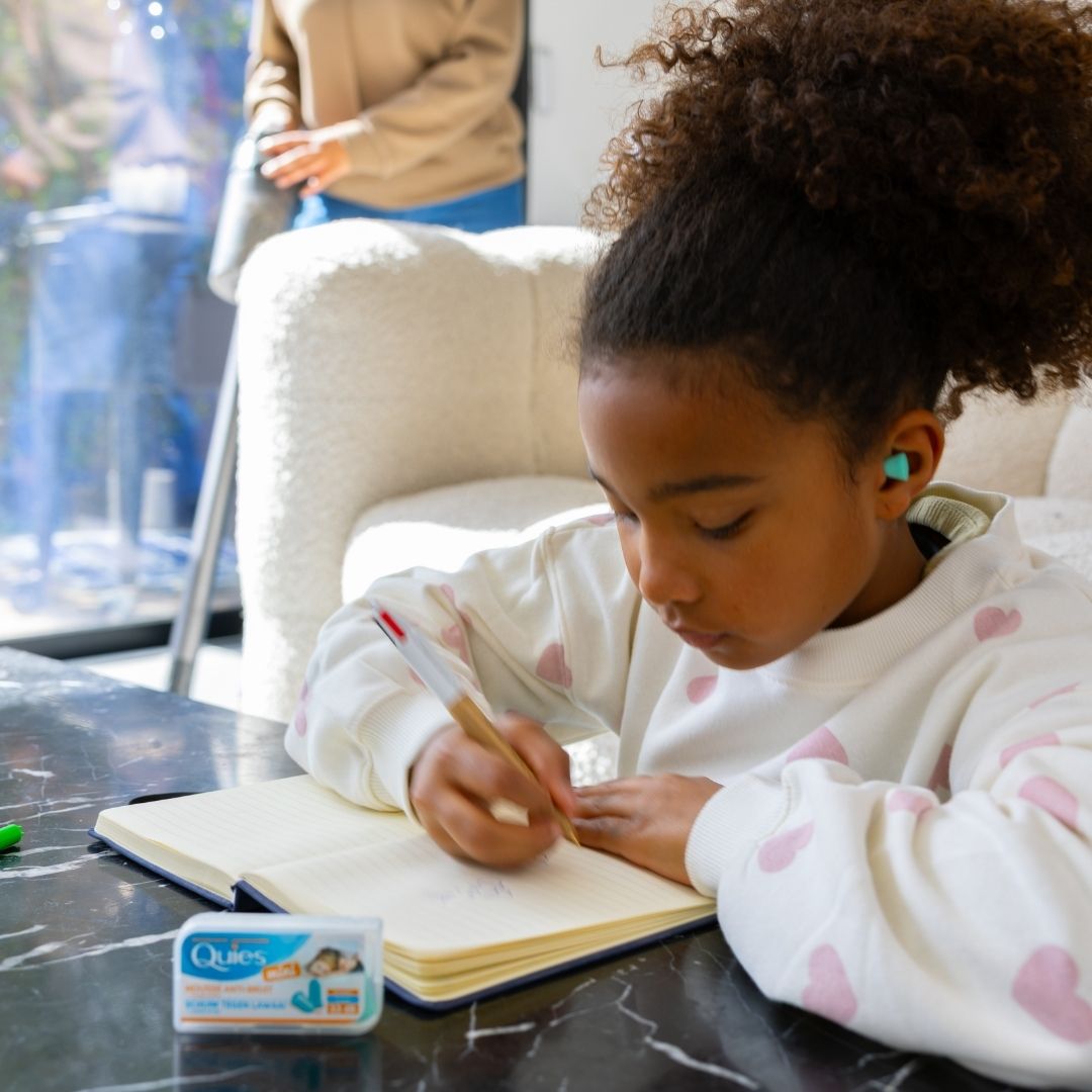Petite fille faisant ses devoirs avec des bouchons d'oreilles Quies Mousse Mini pendant que sa mère passe l'aspirateur
