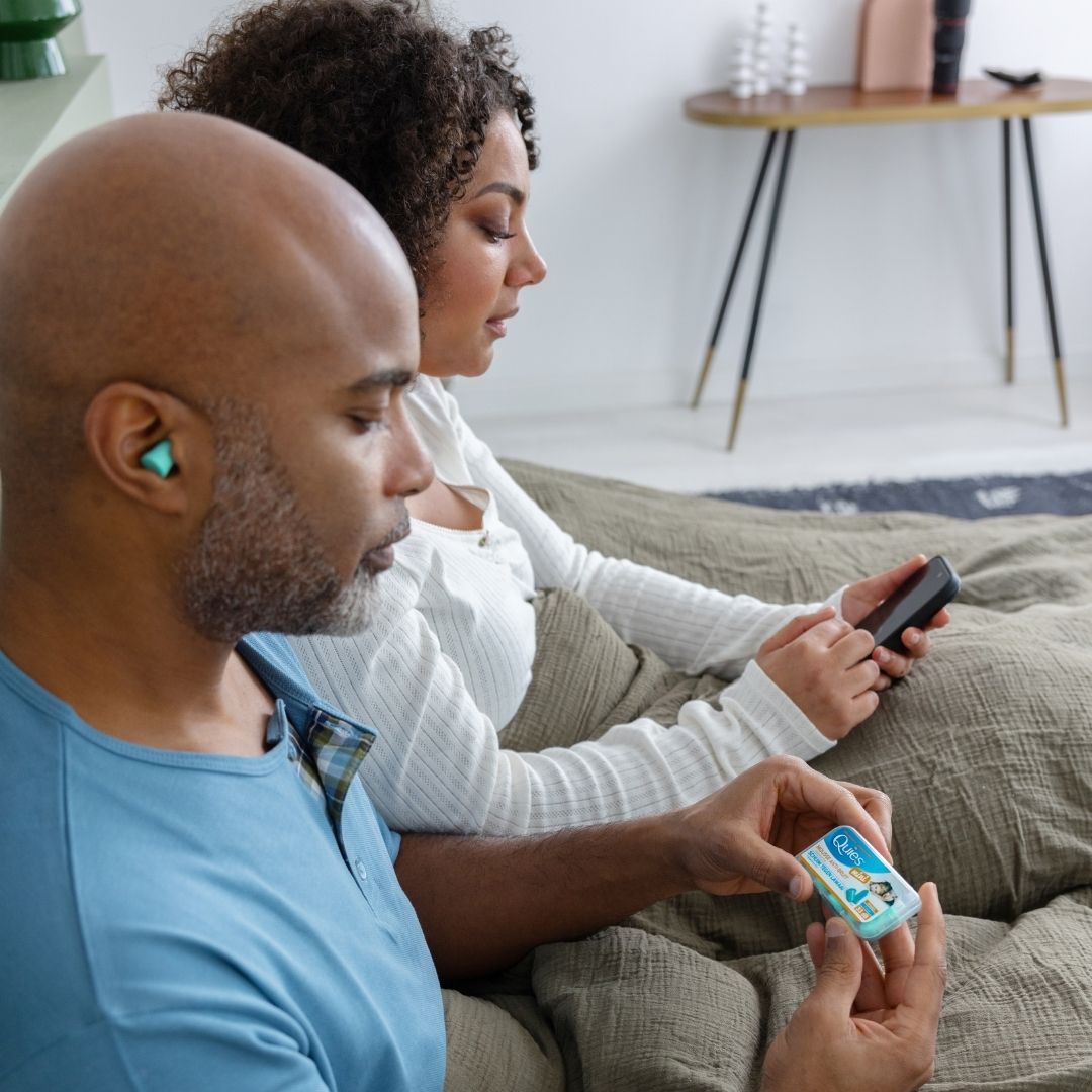 Homme lisant dans son lit avec des bouchons Quies Mousse Mini pendant que sa femme utilise son téléphone